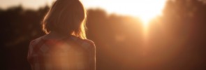 A woman with her back to the camera looks across a field as the sun goes down over distant trees.