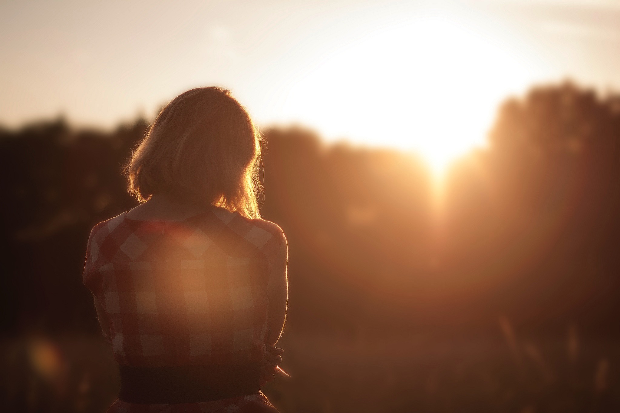 A woman with her back to the camera looks across a field as the sun goes down over distant trees.